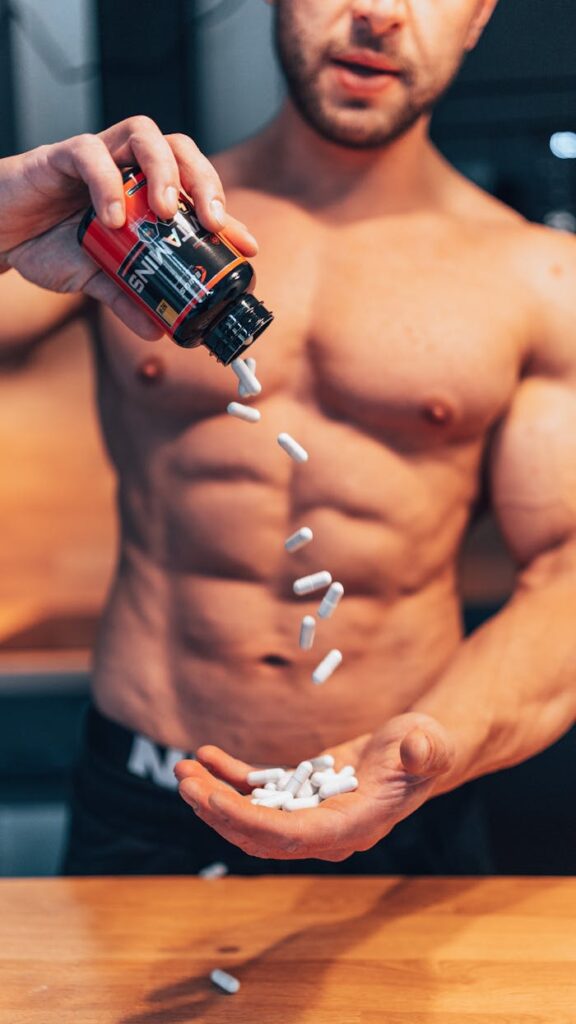 Shirtless man pouring white supplement capsules into his hand with muscles in focus.